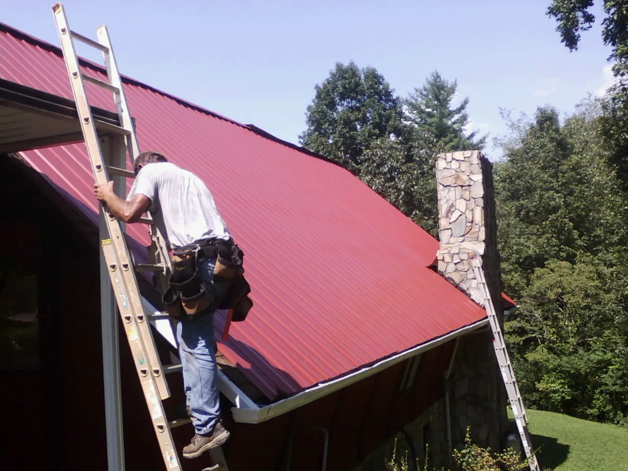 Roofer on a ladder beside a red metal roof and stone chimney — Soffit & Fascia Repair by Roofing Brothers in Lebo